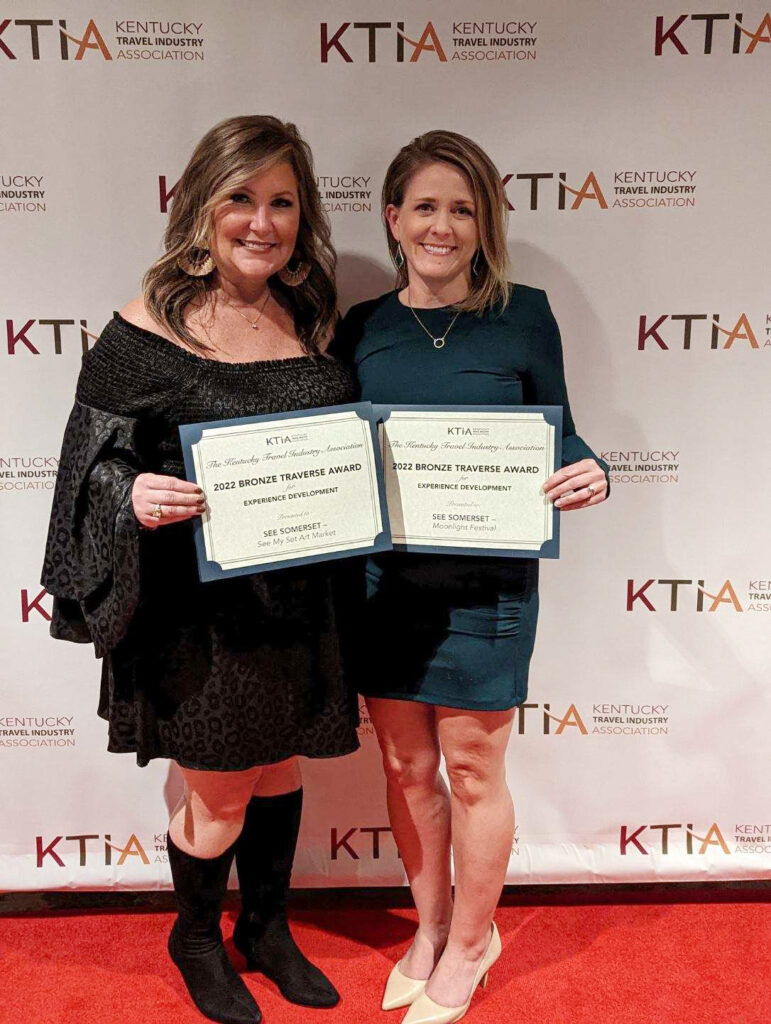 two women holding certificates in front of a step and repeat that says KTIA