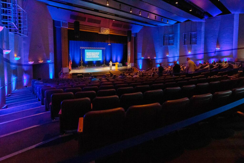 darkly lit auditorium with subdued blue lighting with speaker on stage addressing the audience