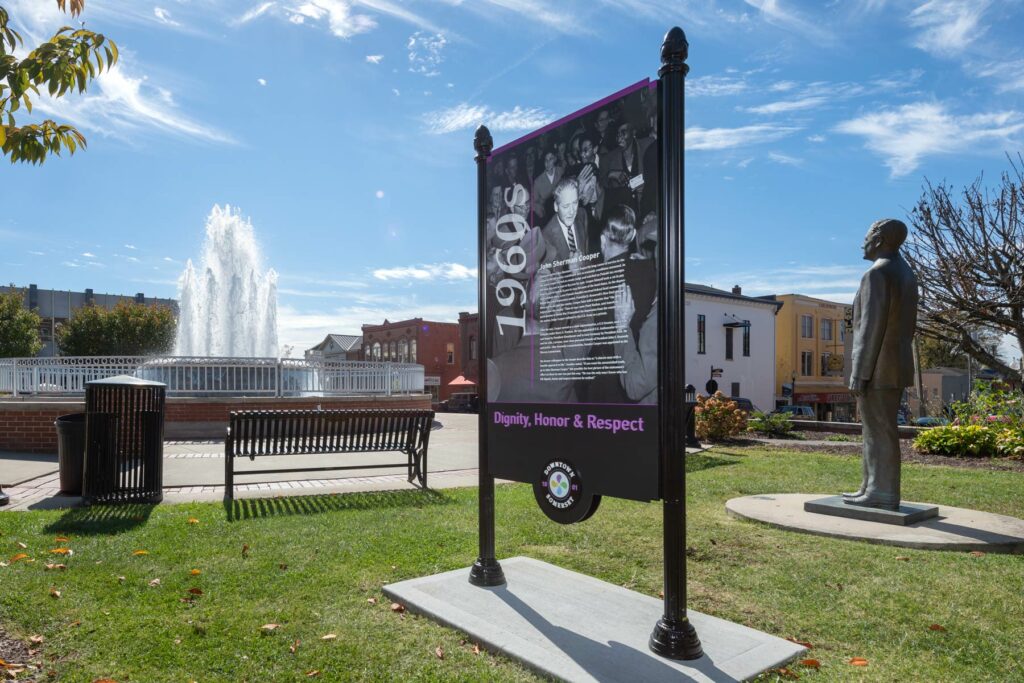 Historic marker sign and statue in park with fountain and buildings in the background