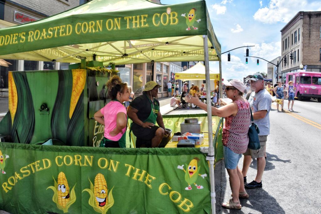 People on the street at a tent that says "Roasted Corn on the Cob"