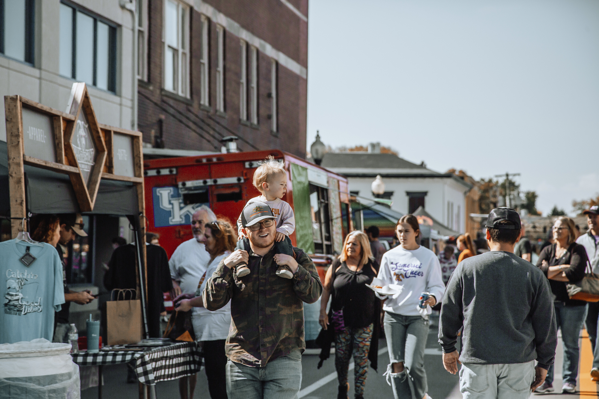 Man walking down street filled with people and foo trucks with a toddler on his shoulders